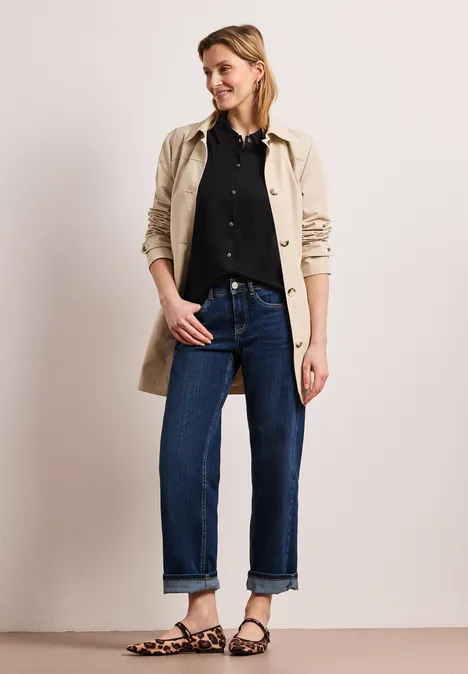 Frau in beiger Trenchcoat, schwarzem Hemd, hochgekrempelten Jeans und Sandalen vor neutralem Hintergrund.